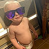 toddler, child, diaper, sunglasses, indoor, air_conditioner, reflection, wooden_table, floor_tiles, bare_chest, person, baby, cute, smiling, standing, home, furniture, lighting, portrait, casual