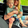 child, woman, car_interior, tattoo, shorts, green_grass, window, seat, seatbelt, armrest, watch, scrunchie, barefoot, pointing, casual_clothing, daylight, portrait, family, toddler, adult