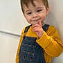 toddler, child, smile, denim_overalls, yellow_shirt, standing, indoor, light_brown_hair, cute, playful, portrait, person, young_child, casual_clothing, wall, expression, gesture, face, happy, one_person