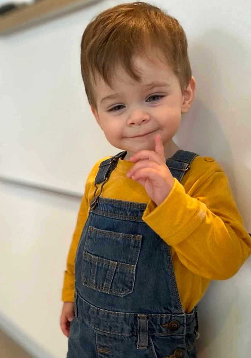 Eyden participe au concours pour gagner de l'argent avec cette photo : toddler, child, smile, denim_overalls, yellow_shirt, standing, indoor, light_brown_hair, cute, playful, portrait, person, young_child, casual_clothing, wall, expression, gesture, face, happy, one_person