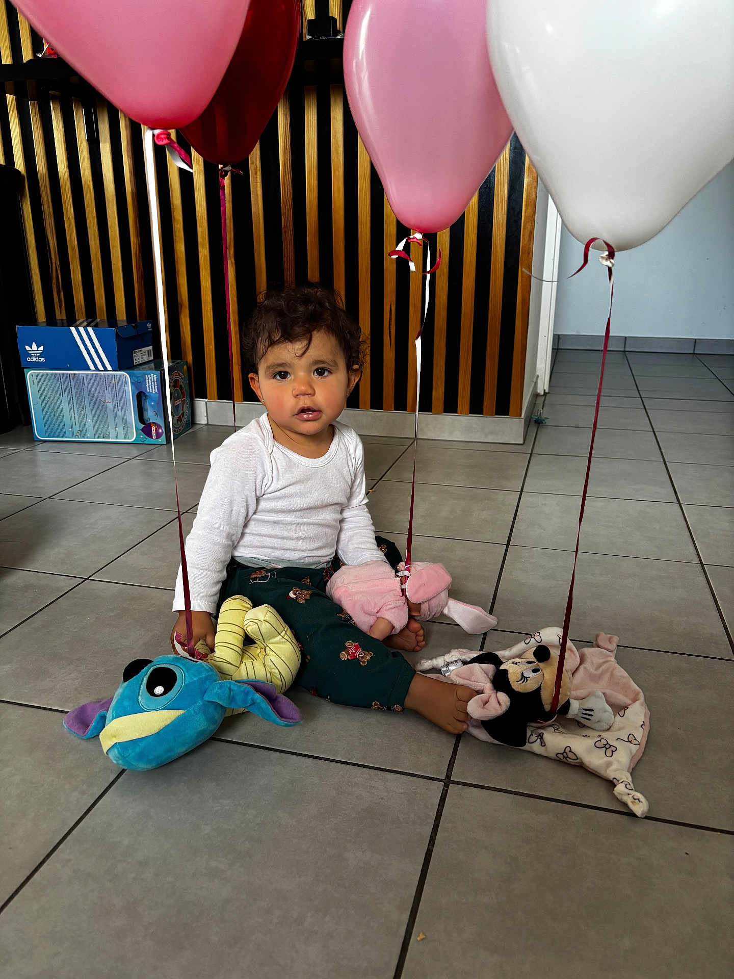 Hayana participe au concours pour gagner de l'argent avec cette photo : toddler, child, balloons, plush_toys, indoor, floor, tiles, white_shirt, pants, curly_hair, brown_eyes, toy_mouse, toy_fish, pink_balloons, white_balloon, sitting, playtime, home, cute, young_child