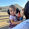 adult, baby, building, caregiver, child, curious, ear_protection, face, fence, hair, headphones, holding, outdoor, person, portrait, shadows, sky, street, sunlight, toddler