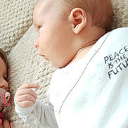 Emiliano a rejoint le concours — aidez-le/la à gagner de superbes lots ! child, baby, toddler, pacifier, onesie, text_on_clothing, sibling, blanket, plush, laying, face, hand, ear, portrait, closeup, cute, bonding, family, soft_texture, indoor