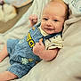 baby, infant, child, denim_overalls, thumbs_up, smile, blanket, soft_texture, cute, portrait, indoors, clothing, relaxed, happy, face, expression, skin, head, arm, person