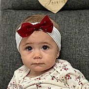 Madison is registered to the contest to win money with this photo: baby, headband, red_bow, floral_clothing, couch, gray_couch, heart_shaped_sign, wooden_sign, indoor, portrait, child, infant, cute, serious_expression, sitting, young_child, face, person, head, clothing