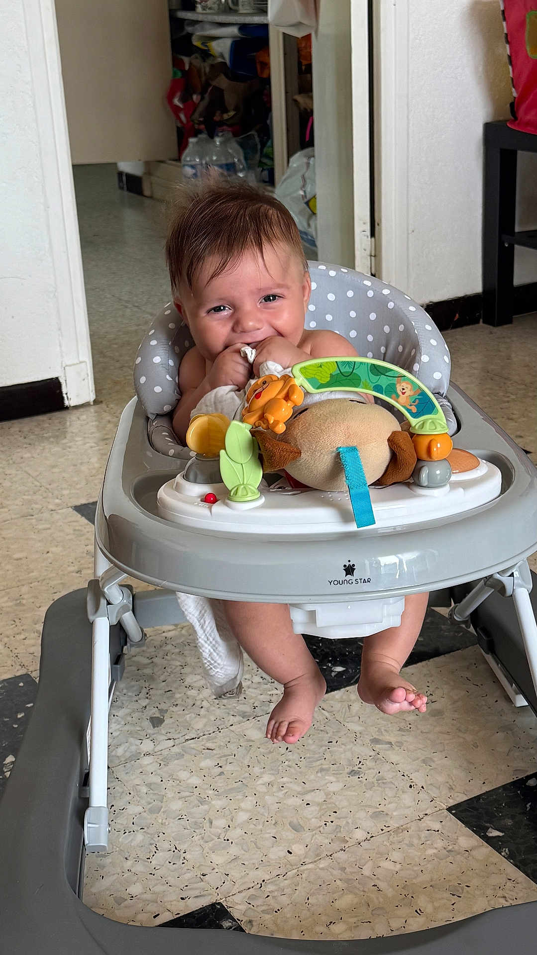 Jules participe au concours pour gagner de l'argent avec cette photo : baby, walker, toy, plush_toy, floor, tile_floor, indoor, child, smiling, chewing, clothing, foot, seat, furniture, polka_dot, happy, person, home, doorway, playtime