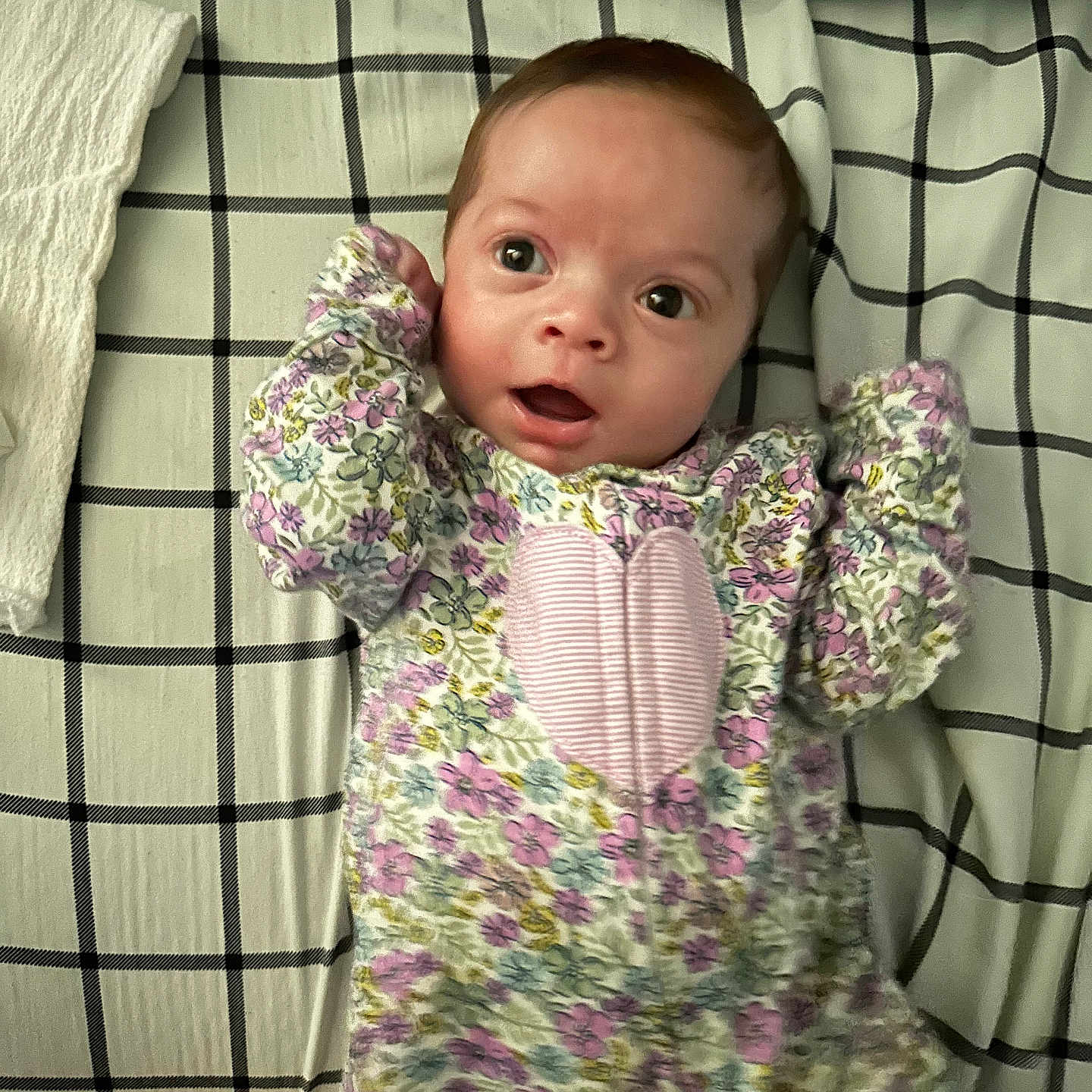 Evelynn is registered to the contest to win money with this photo: baby, bedspread, checkered_pattern, child, curious_expression, cute, fabric_texture, face, floral_onesie, hand_up, head, indoors, infant, lying_down, pink_heart, portrait, sleepwear, soft_lighting, wide_eyes, young_child