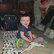 Blake is registered to the contest to win money with this photo: adult, baby, bag, blanket, carpet, casual, child, clothing, couch, cup, furniture, indoor, living_room, person, play, slippers, smile, toddler, toy, wall_photos
