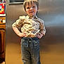 appliance, boy, casual_clothing, child, cute, dinosaurs, hands_on_hips, happy, home, indoor, jeans, kitchen, person, portrait, refrigerator, shirt, smiling, standing, wooden_floor, young