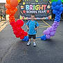 child, boy, balloon_arch, rainbow_colors, school, backpack, outdoor, banner, celebration, smiling, asphalt, sunlight, shorts, sneakers, striped_shirt, people, parking_lot, daylight, event, happy