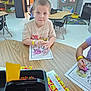 art_supplies, child, classroom, coloring, coloring_activity, crayons, education, furniture, indoor, kids, learning, pencils, school, seated, smiley_face_labels, table, wooden_table, worksheet, writing_tools, young_child