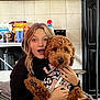 Oakley joined the competition — help win amazing prizes! person, dog, indoor, cuddling, curly_hair, black_sweater, graphic_shirt, brown_dog, kitchen, snacks, drinks, counter, tile_wall, surprised_expression, pet, cozy, furniture, blanket, table, home