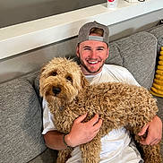 Oakley is registered to the contest to win money with this photo: man, dog, couch, smile, cap, curly_hair, white_shirt, bracelet, decor, owl_figurine, drink_cup, indoor, happy, pet, furniture, casual, seated, relaxed, home, friendship