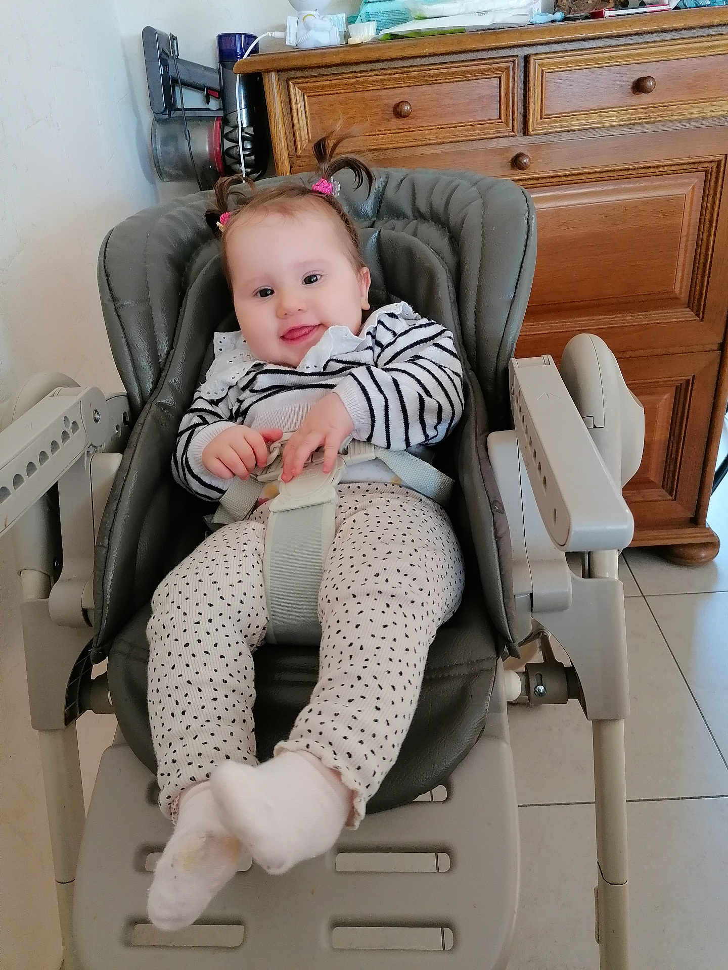 Charline a rejoint le concours — aidez-le/la à gagner de superbes lots ! baby, child, high_chair, pigtails, striped_sweater, polka_dot_pants, socks, smiling, indoor, wooden_cabinet, floor_tiles, seat_belt, baby_seat, face, person, cute, happy, hair_ties, furniture, tongue_out