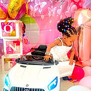 Ifunanya participe au concours pour gagner de l'argent avec cette photo : african_american_child, balloons, birthday_party, butterflies, celebration, child_playing, colorful_hair_clips, cute, decorations, event, floor, happy, indoor, party, pink_backdrop, small_car, toddler, toy_car, watch, white_dress