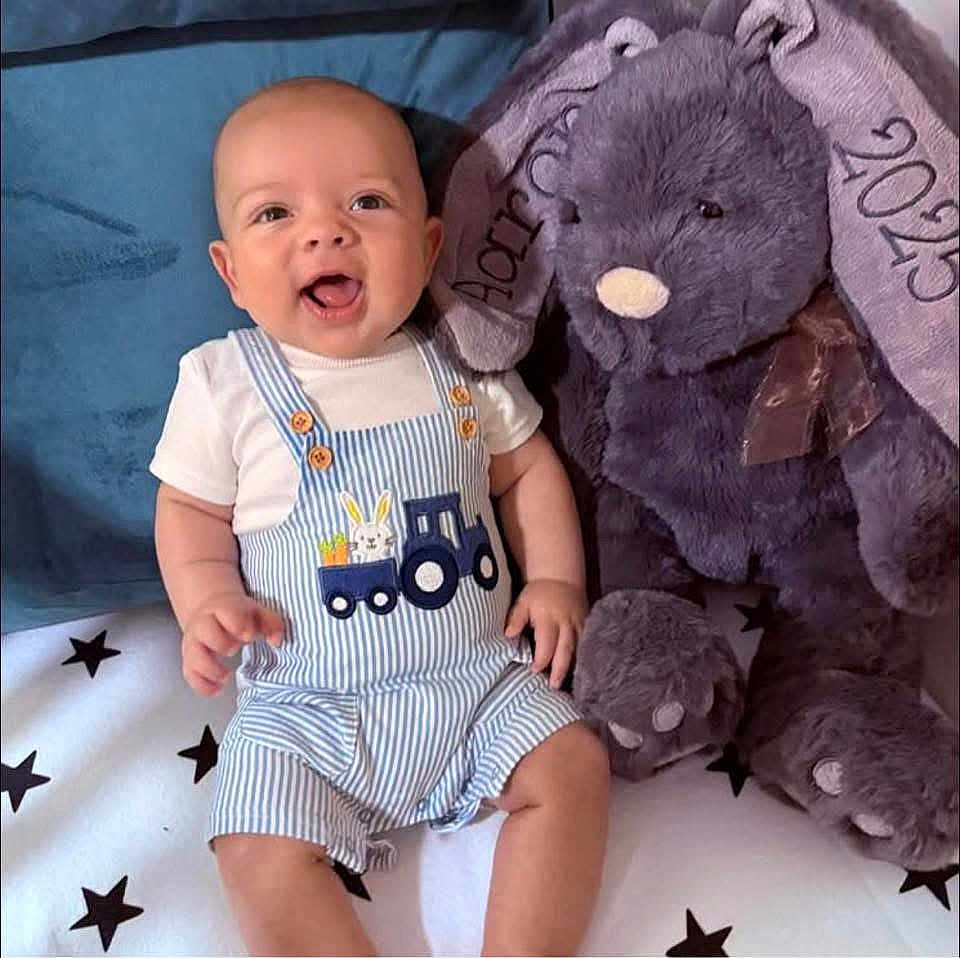 Aaron is registered to the contest to win money with this photo: baby, child, smiling, overalls, striped_clothing, white_tshirt, plush_toy, teddy_bear, blanket, stars, sitting, cute, happy, indoor, soft_toy, ribbon, embroidery, infant, person, face