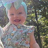 Jaina is registered to the contest to win money with this photo: baby, bow, child, cute, daylight, floral, greenery, happy, headband, holding, lens_flare, nature, outdoor, person, portrait, romper, smiling, summer, sunlight, trees