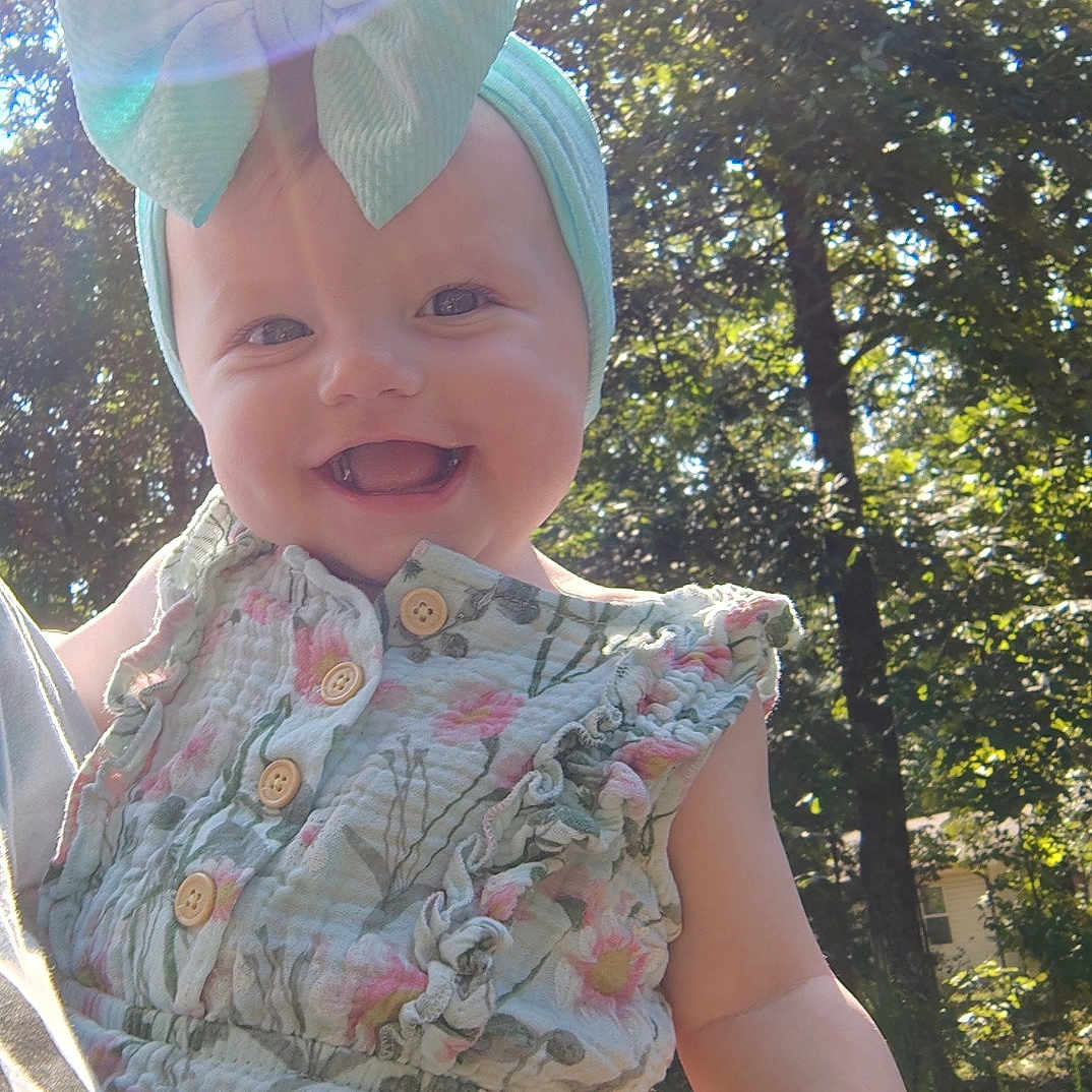 Jaina is registered to the contest to win money with this photo: baby, bow, child, cute, daylight, floral, greenery, happy, headband, holding, lens_flare, nature, outdoor, person, portrait, romper, smiling, summer, sunlight, trees
