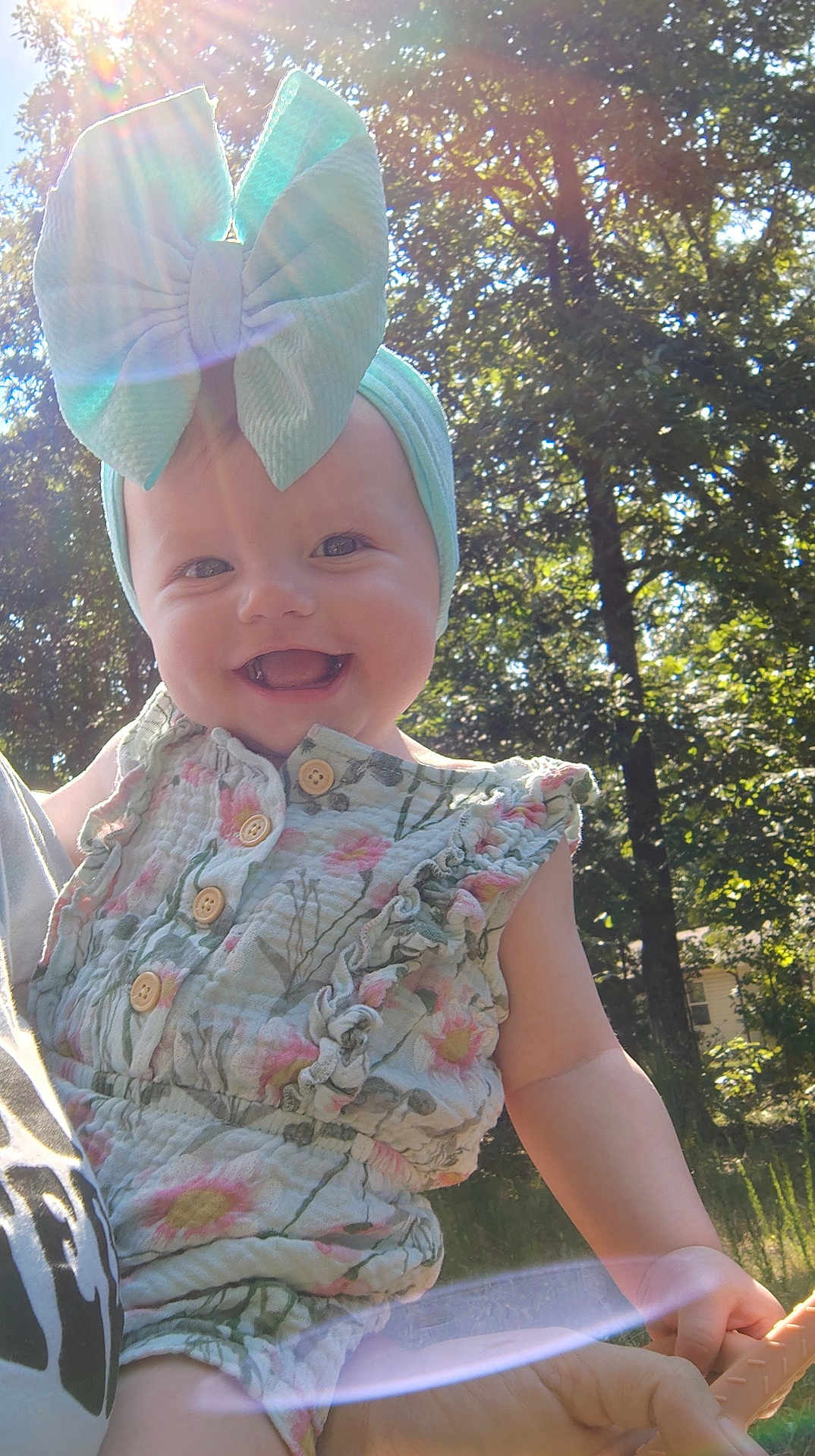 Jaina is registered to the contest to win money with this photo: baby, bow, child, cute, daylight, floral, greenery, happy, headband, holding, lens_flare, nature, outdoor, person, portrait, romper, smiling, summer, sunlight, trees