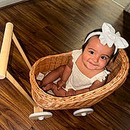 Beatriz joined the competition — help win amazing prizes! baby, child, wicker_basket, bow, headband, smile, wooden_floor, toy_cart, happy, indoors, cute, sitting, person, infant, playful, white_clothing, flooring, basket, wheel, portrait