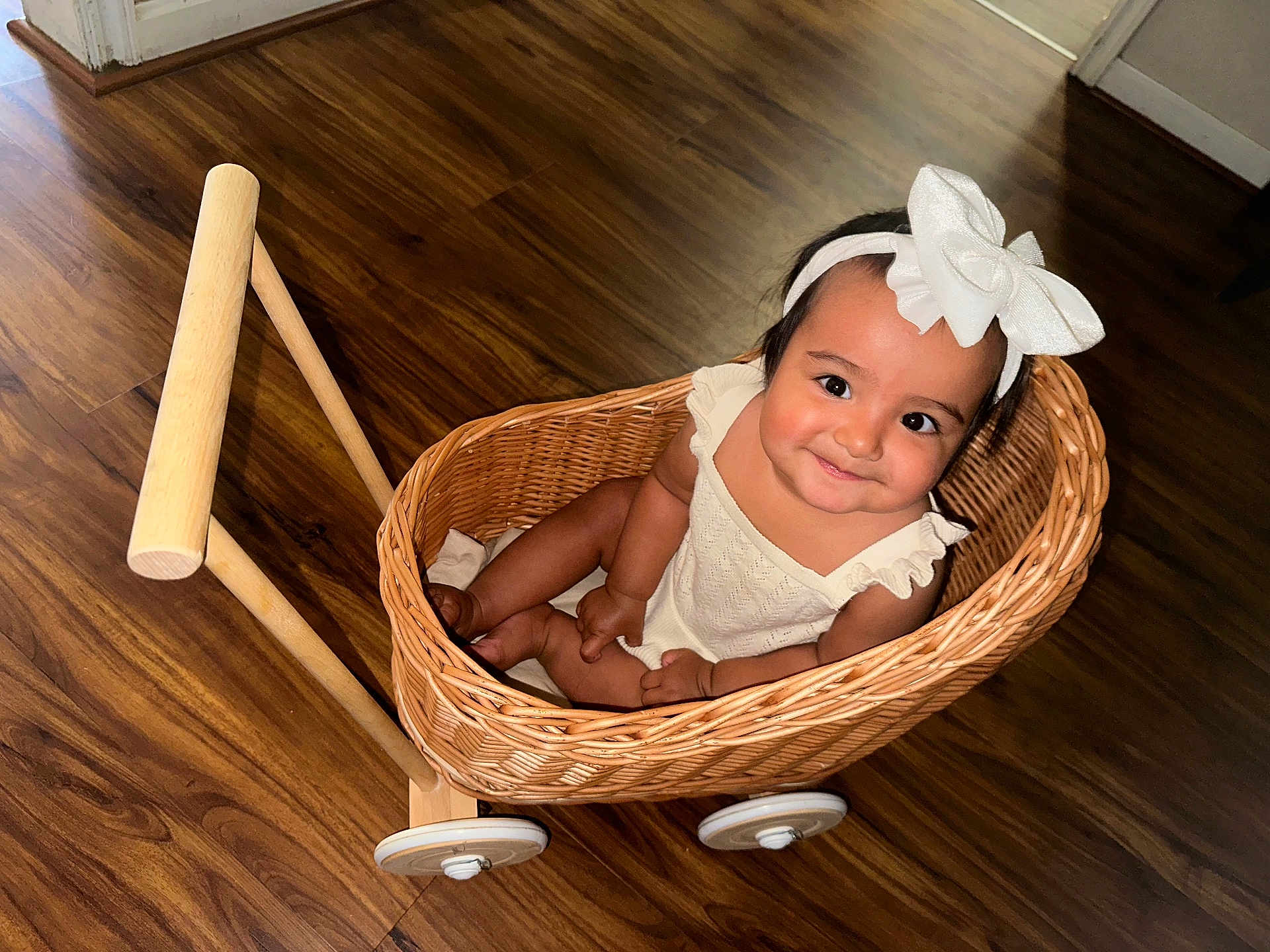 Beatriz joined the competition — help win amazing prizes! baby, child, wicker_basket, bow, headband, smile, wooden_floor, toy_cart, happy, indoors, cute, sitting, person, infant, playful, white_clothing, flooring, basket, wheel, portrait
