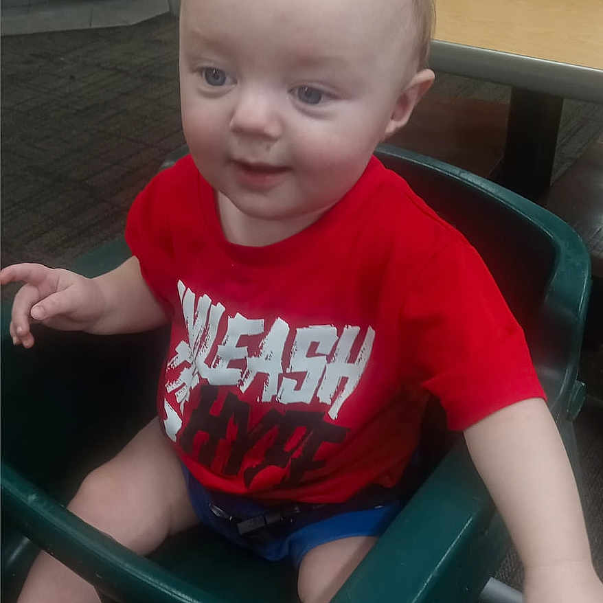 Grayson is registered to the contest to win money with this photo: baby, bodypart, chair, clothing, diningtable, e_scooter, face, furniture, happy, head, person, photography, portrait, shorts, sitting, smile, t_shirt, table, transportation, vehicle