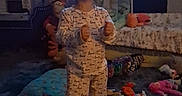 Nova is registered to the contest to win money with this photo: child, toddler, smiling, happy, standing, pajamas, indoors, bedroom, toys, stuffed_animals, carpet, couch, pillows, television, portrait, full_body, thumbs_up, playroom, dim_lighting, home