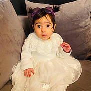 Rehmat is registered to the contest to win money with this photo: baby, child, pigtails, white_dress, lace, sunglasses, purple_sunglasses, couch, gray_couch, indoor, sitting, wide_eyes, cute, portrait, infant, expression, clothing, cozy, young_child, person
