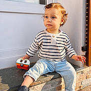 Côme participe au concours pour gagner de l'argent avec cette photo : toddler, child, curly_hair, striped_shirt, light_blue_jeans, toy, camper_toy, navy_shoes, outdoor, stone_ledge, sunlight, window, door, casual_clothing, sitting, curious, young_child, daylight, expression, portrait
