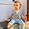 toddler, child, curly_hair, striped_shirt, light_blue_jeans, toy, camper_toy, navy_shoes, outdoor, stone_ledge, sunlight, window, door, casual_clothing, sitting, curious, young_child, daylight, expression, portrait