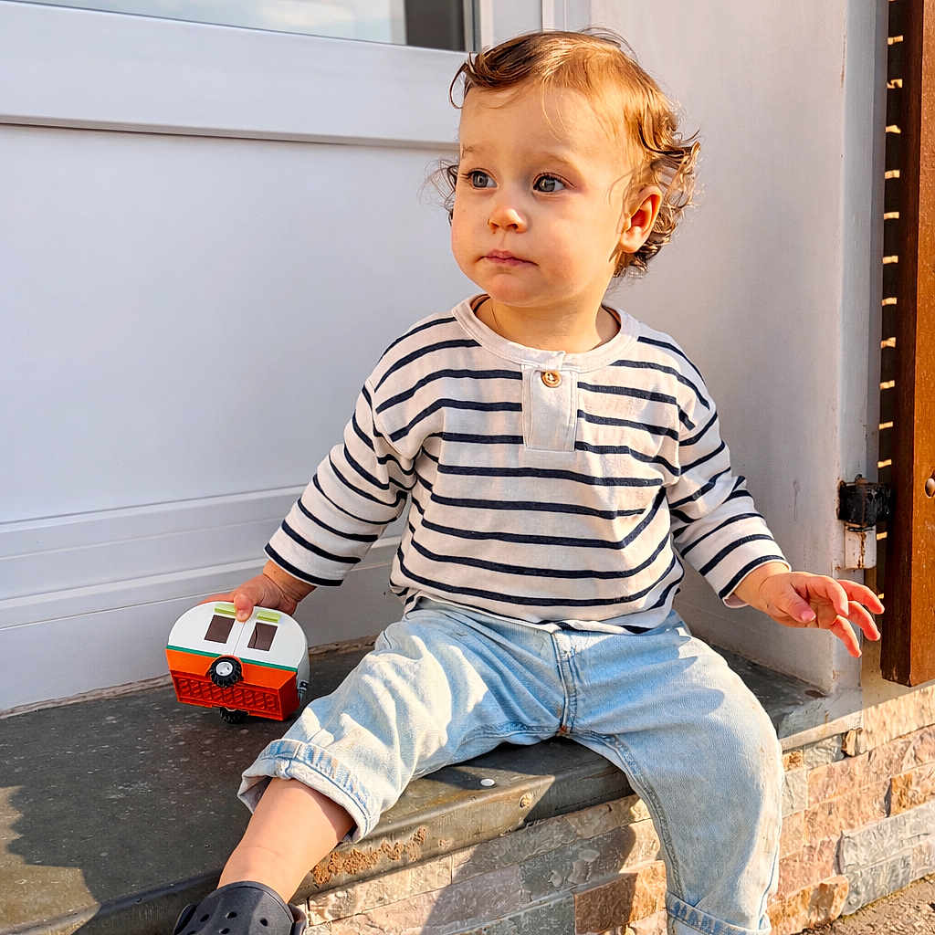 Côme participe au concours pour gagner de l'argent avec cette photo : toddler, child, curly_hair, striped_shirt, light_blue_jeans, toy, camper_toy, navy_shoes, outdoor, stone_ledge, sunlight, window, door, casual_clothing, sitting, curious, young_child, daylight, expression, portrait