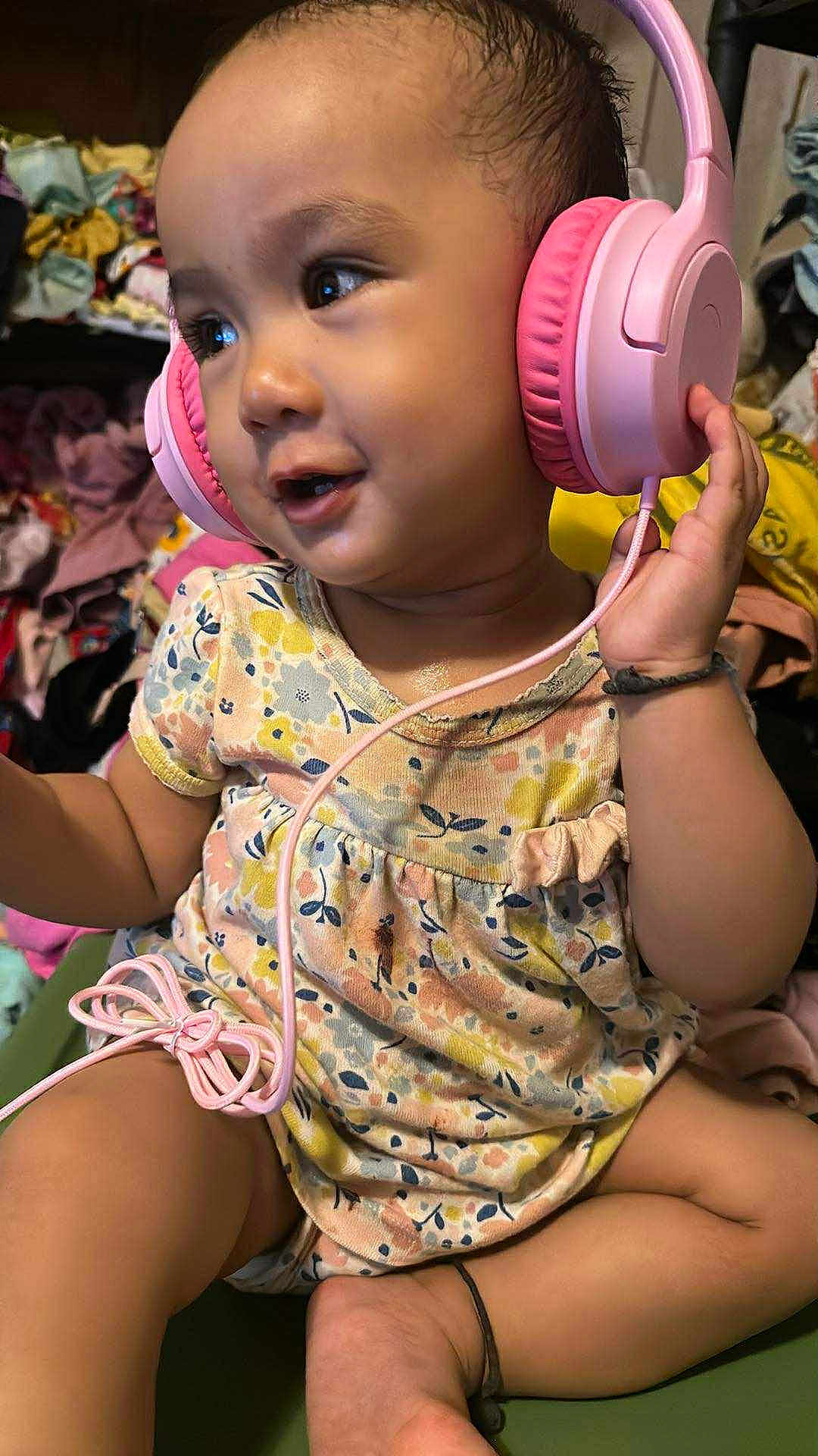 Sara is registered to the contest to win money with this photo: toddler, child, headphones, pink_headphones, floral_dress, sitting, indoors, happy, curious, clothing, baby, person, music, technology, cute, portrait, skin, expression, hand, foot