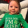 baby, infant, smile, green_clothing, onesie, text, shamrock, face, hair, blanket, red_background, cozy, portrait, cute, child, happy, indoors, person, sitting, clothing