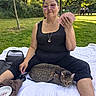 woman, cat, playing_cards, park, blanket, glasses, trees, grass, outdoor, sitting, leisure, pet, harness, picnic, people, bench, sunlight, casual_clothing, relaxation, nature