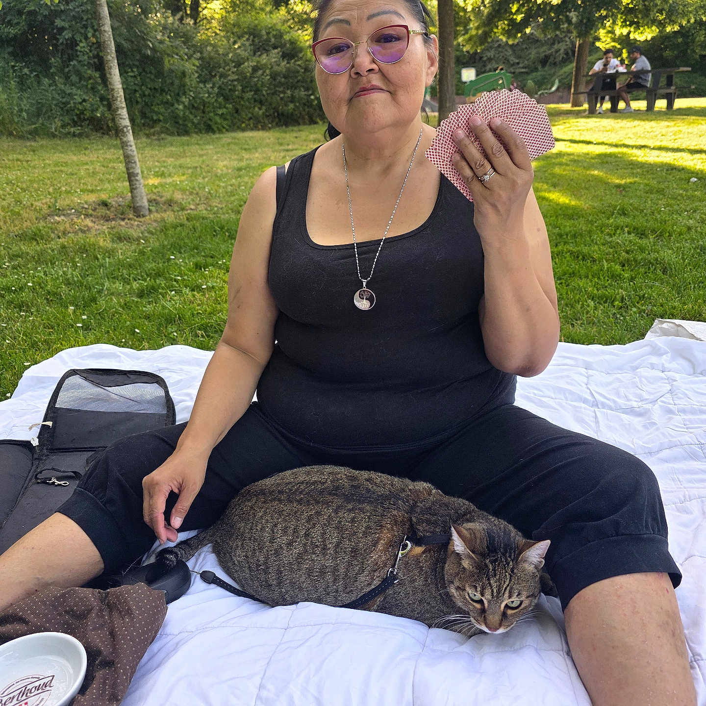 Chipinette participe au concours pour gagner de l'argent avec cette photo : woman, cat, playing_cards, park, blanket, glasses, trees, grass, outdoor, sitting, leisure, pet, harness, picnic, people, bench, sunlight, casual_clothing, relaxation, nature
