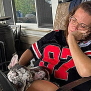 Birdie is registered to the contest to win money with this photo: woman, sleeping, dog, french_bulldog, pet, chair, jersey, glasses, porch, window, outdoor, relaxing, nap, black_clothing, red_numbers, furniture, hand, resting, calm, domestic