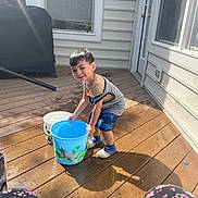 Benjamin is registered to the contest to win money with this photo: child, boy, bucket, water, deck, outdoor, sunlight, smiling, clothing, house, window, door, floor, shadow, play, person, summer, fun, legs, patterned_clothing