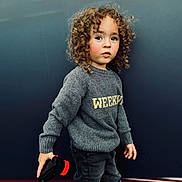 Ibrahim participe au concours pour gagner de l'argent avec cette photo : casual_clothing, child, color_contrast, curly_hair, fashion, gray_sweater, jeans, looking_away, outdoor, person, playful, portrait, serious_expression, short_hair, standing, street_style, toddler, toy, urban_background, young_child