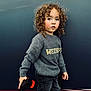 casual_clothing, child, color_contrast, curly_hair, fashion, gray_sweater, jeans, looking_away, outdoor, person, playful, portrait, serious_expression, short_hair, standing, street_style, toddler, toy, urban_background, young_child