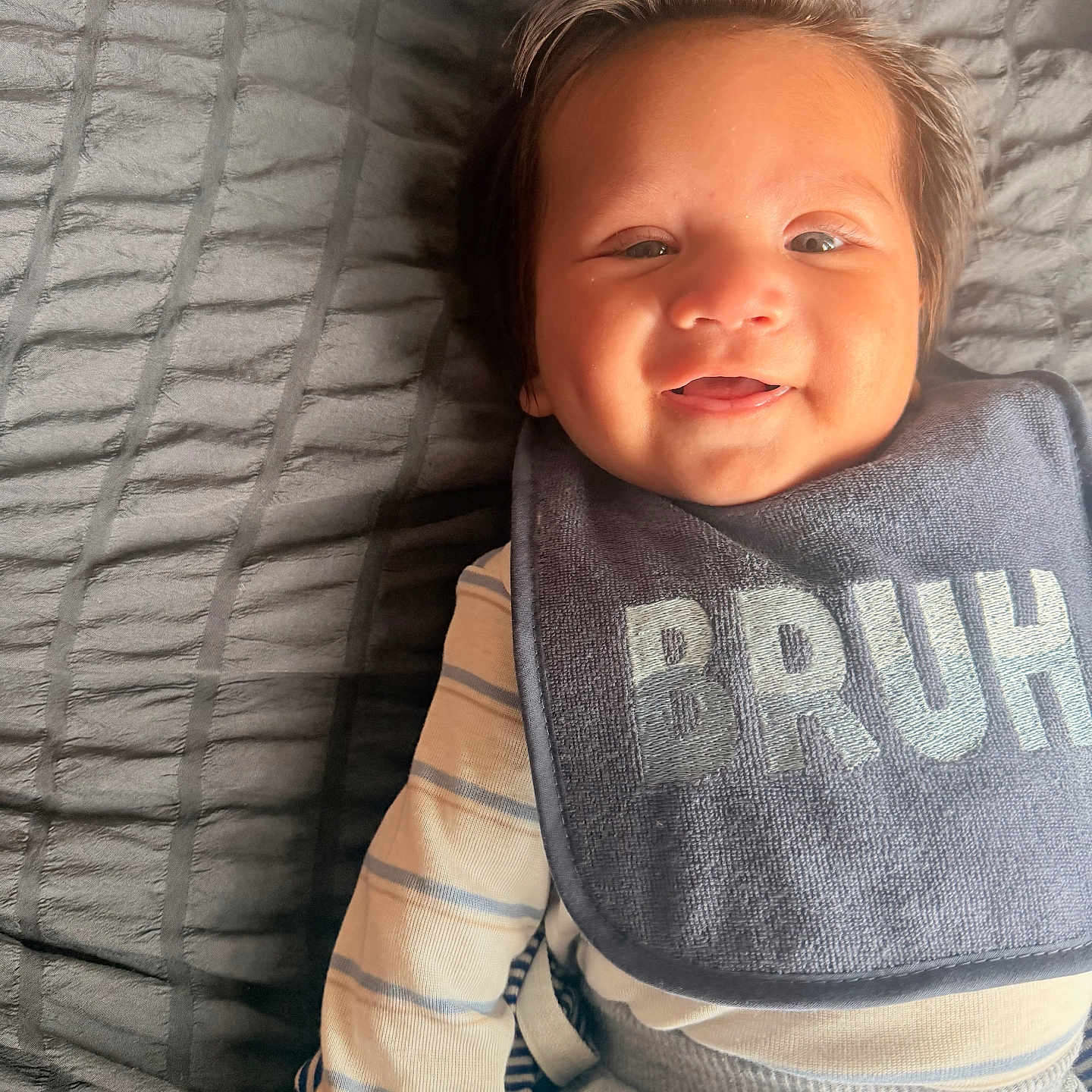 Jesus is registered to the contest to win money with this photo: baby, smile, bib, clothing, striped_shirt, gray_blanket, hair, happy, infant, portrait, lying_down, cute, child, face, toddler, textile, fabric, indoor, person, soft