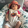 Jesus joined the competition — help win amazing prizes! baby, child, hat, bucket_hat, romper, elmo, cushion, leaf_pattern, indoor, seated, curious, face, clothing, person, infant, cute, red, beige, comfortable, portrait