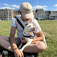 Peppa participe au concours pour gagner de l'argent avec cette photo : affection, blue_sky, buildings, cap, casual, clouds, daytime, dog, grass, harness, leash, man, outdoor, park, pet, relaxed, shorts, sitting, sunny, white_tshirt