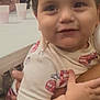 arm, baby, casual, child, close_up, cup, curly_hair, face, fire_truck, holding, indoor, people, person, red, shirt, smiling, striped_pants, table, toddler, white