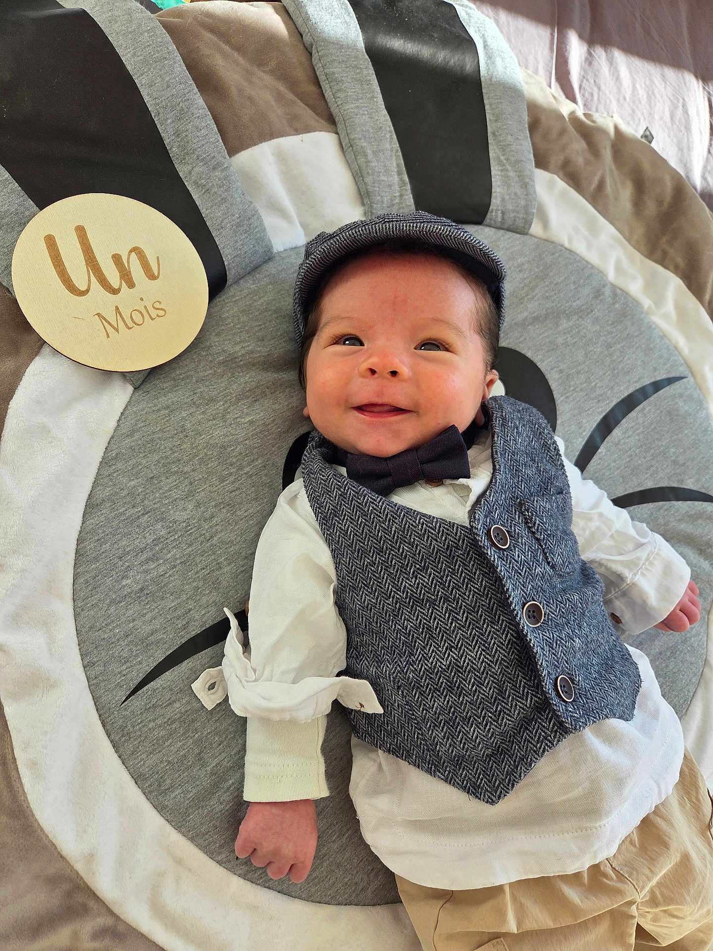 Lowen participe au concours pour gagner de l'argent avec cette photo : baby, infant, smiling, face, vest, bow_tie, cap, play_mat, wooden_plaque, clothing, happy, lying_down, one_month, cute, child, indoor, portrait, fabric, buttoned_vest, infant_boy