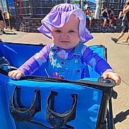 Calliope is registered to the contest to win money with this photo: baby, child, purple_hat, blue_stroller, outdoor, sunny, people, concrete, fence, casual_clothing, daylight, hat, person, purple_shirt, denim_overalls, expression, grip, summer, background, recreation_area