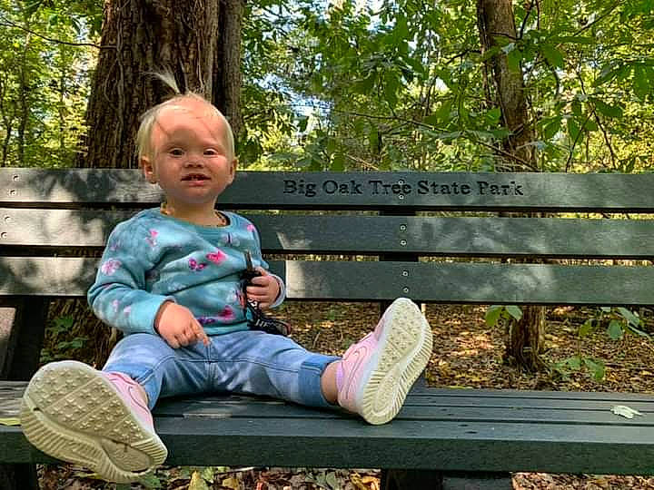 Dixie is registered to the contest to win money with this photo: baby_toddler_clothing, bench, botany, garden, grass, happy, leaf, leg, leisure, outdoor_bench, outdoor_furniture, people_in_nature, person, plant, public_space, recreation, sitting, spring, toddler, tree