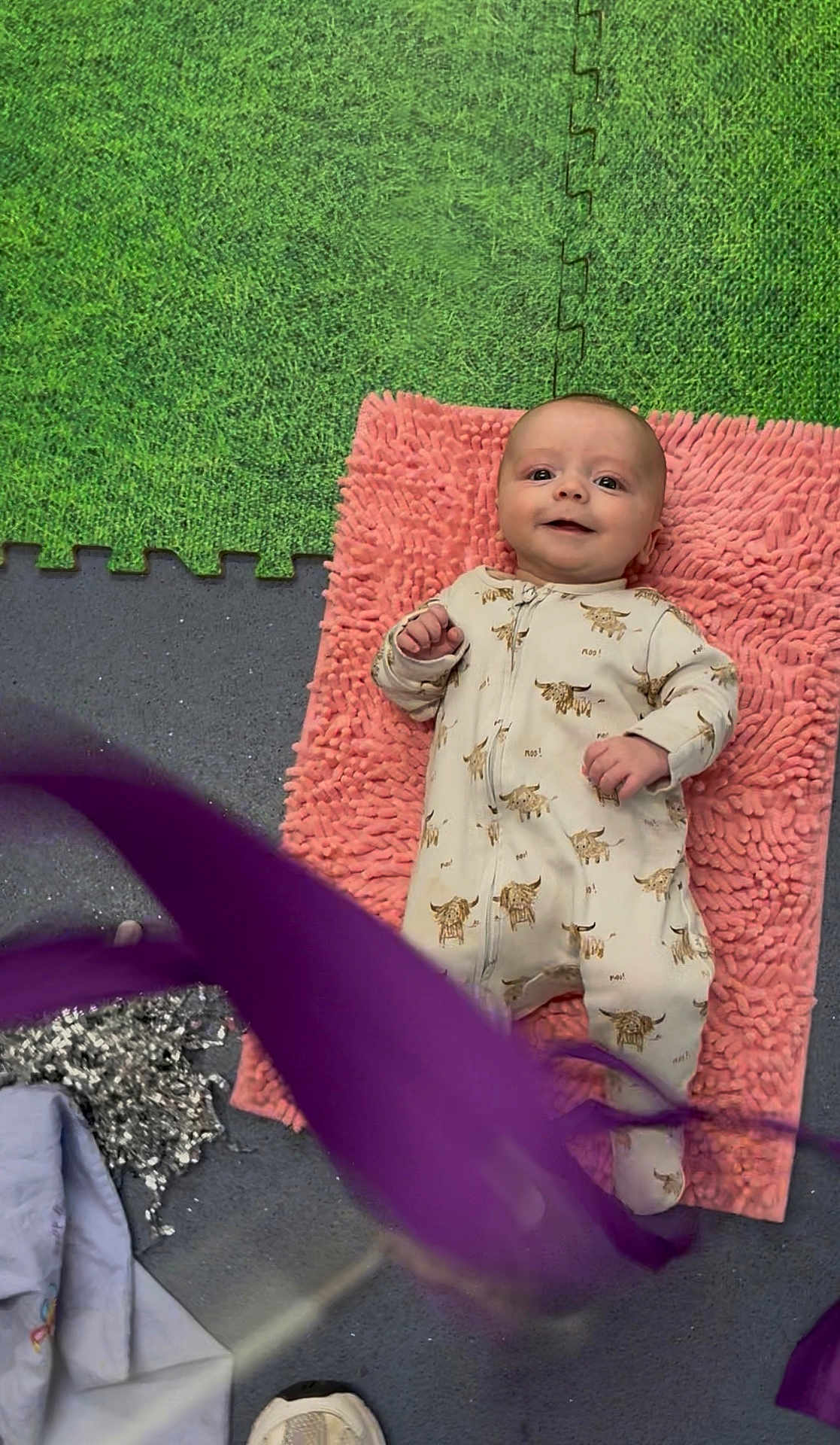 Reggie is registered to the contest to win money with this photo: baby, infant, smiling, onesie, pink_mat, green_floor, foam_tiles, playful, cute, lying_down, child, floor, texture, animal_print, purple_object, footwear, blanket, indoor, happy, portrait