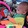 baby, child, doll, car_interior, hand, steering_wheel, parking_permit, green_clothing, red_hair, toy, seat, sunlight, shadow, hand_support, car_window, parking_lot, vehicle, fabric, colorful, person