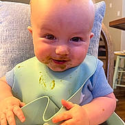 Levi joined the competition — help win amazing prizes! baby, child, face, messy, smile, high_chair, bib, blue_clothing, hands, indoor, home, furniture, wooden_chair, table, cushion, skin, infant, cute, portrait, feeding