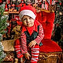 child, christmas, holiday, elf_costume, red_hat, striped_clothing, red_chair, wooden_floor, nutcracker, christmas_tree, decorations, gift_boxes, festive, boots, toy, holiday_season, portrait, indoor, smiling, cute